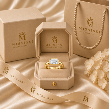Serein east-west emerald-cut bezel engagement ring in 14K yellow gold, presented in coordinated champagne-beige MISSISSI packaging.